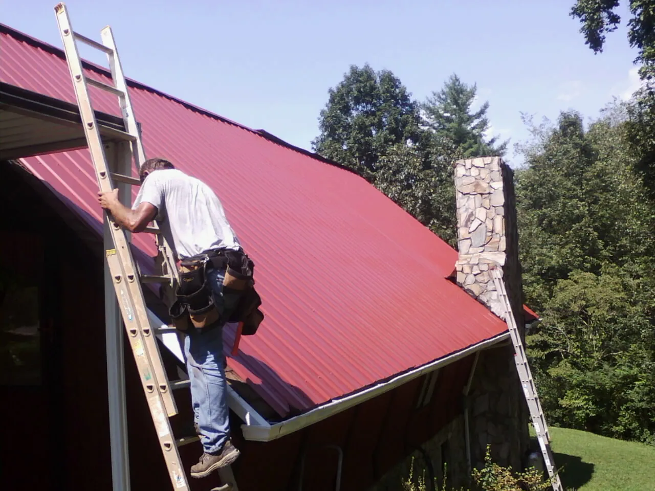Roofer on a ladder beside a red metal roof and stone chimney — Soffit & Fascia Repair by Roofing Brothers in Summerhill